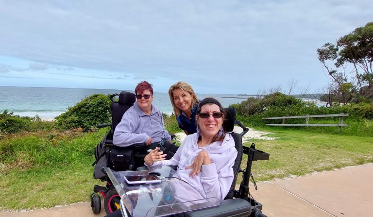 3 people, 2 in wheelchairs, posing for photo next to beach